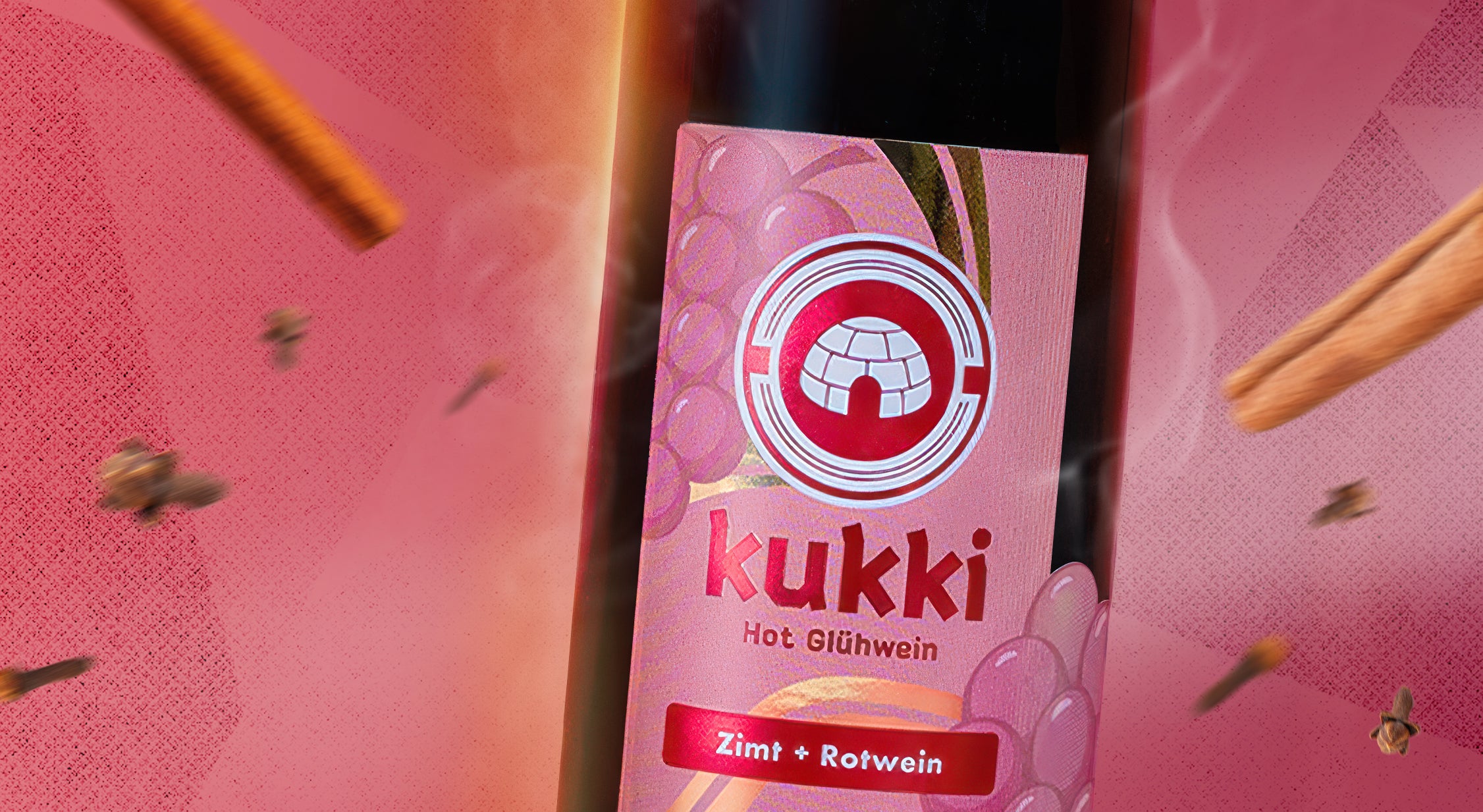 Flasche kukki Cocktail Hot Glühwein mit rosa Etikett, umgeben von Zimtstangen und Nelken vor pinkem Hintergrund.