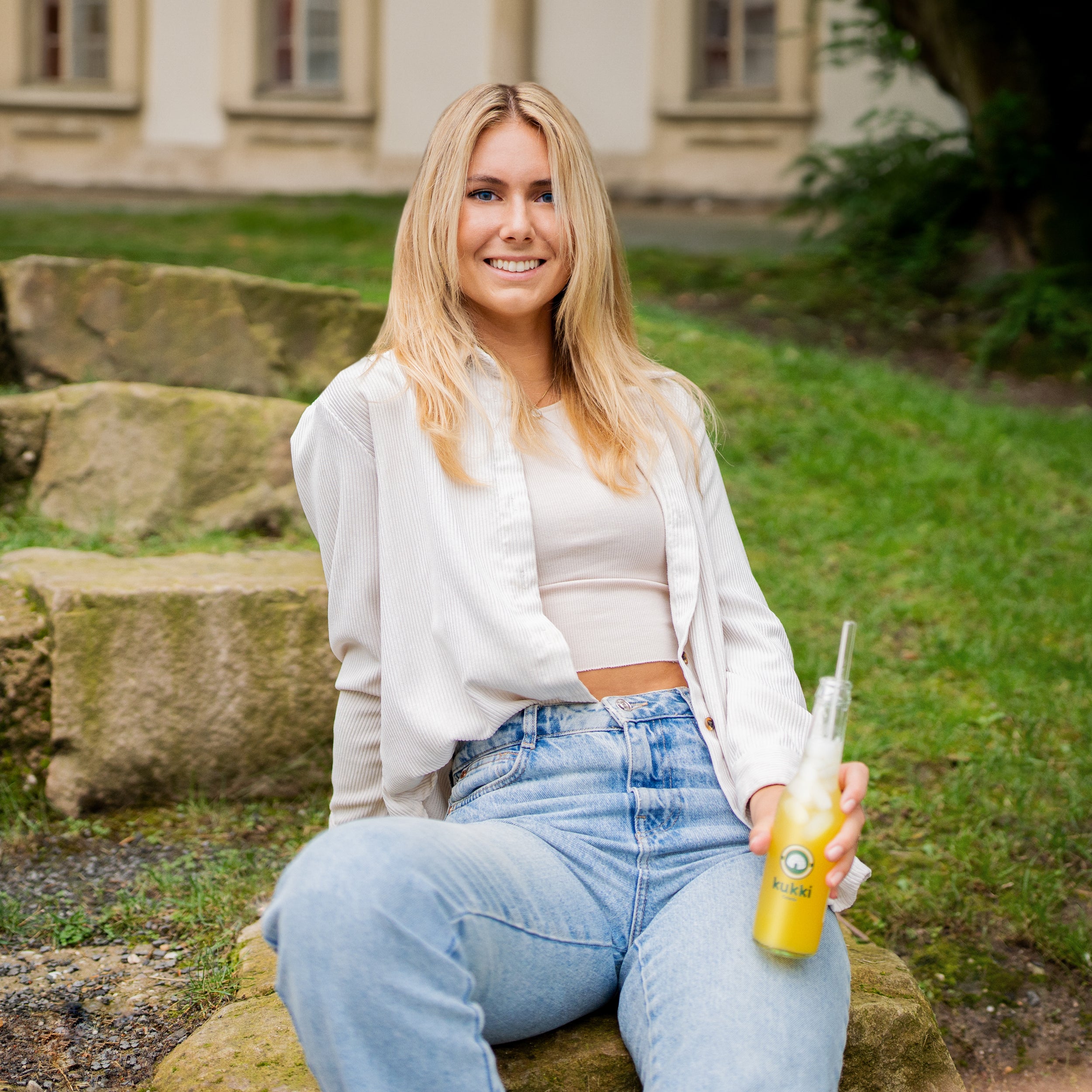 Junge blonde Frau sitzt im Freien und hält eine Glasflasche kukki Cocktail mit gelb-orangefarbenem Getränk.