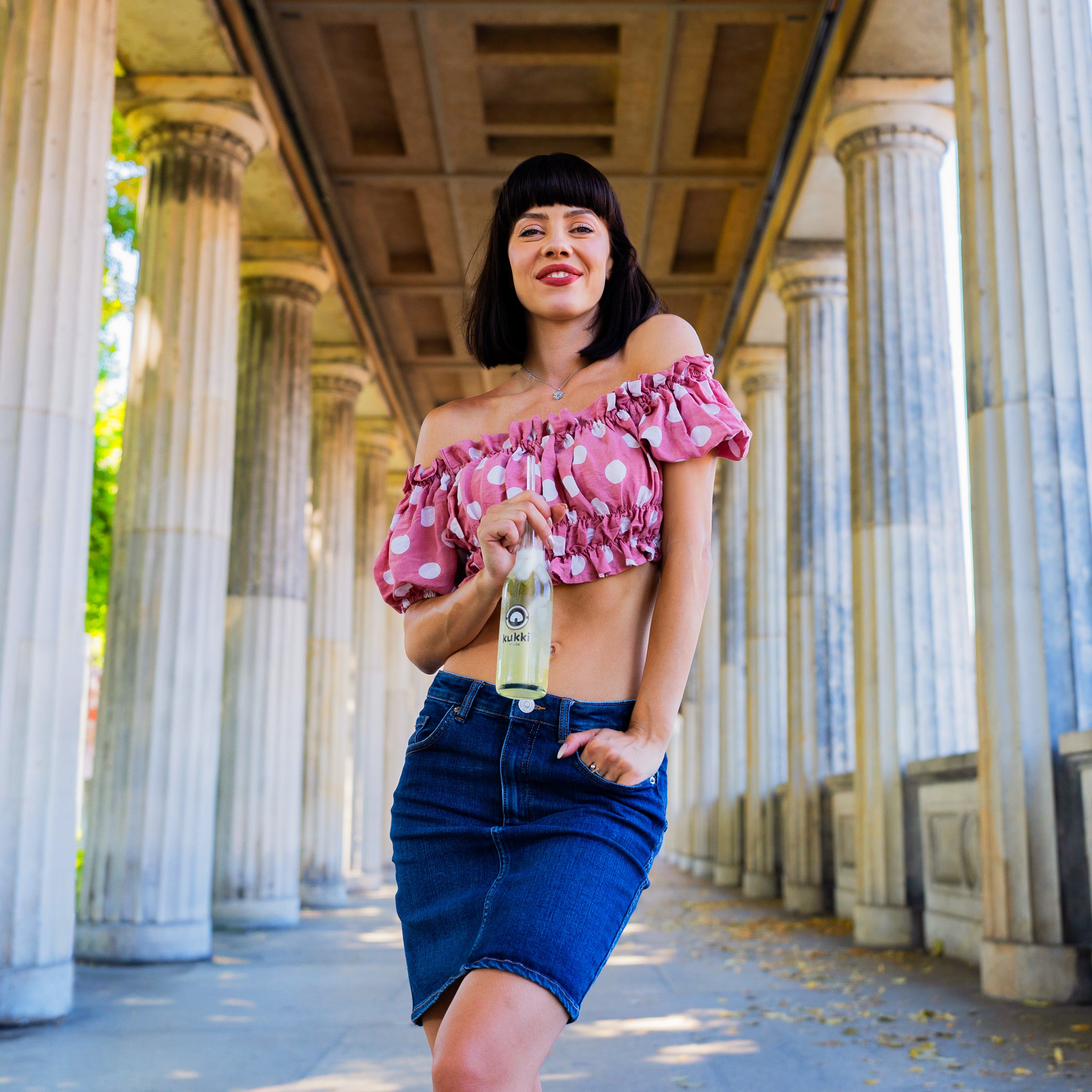 Junge Frau im gepunkteten Crop-Top und Jeansrock hält kukki Cocktail-Flasche im Freien.