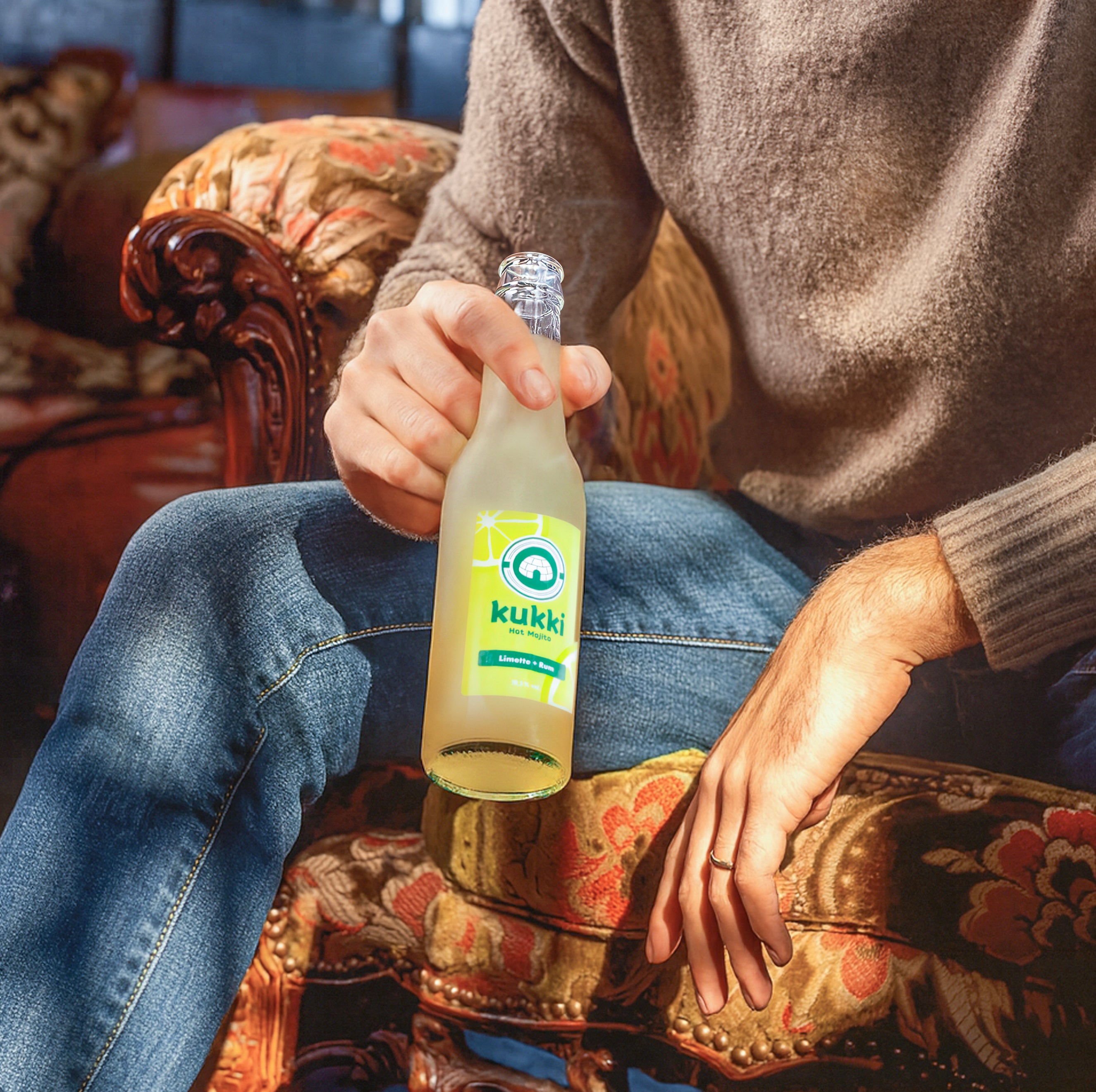 Hand hält gefrostete Glasflasche kukki Cocktail Mojito mit Limette vor gemustertem Sessel in warmer Atmosphäre.
