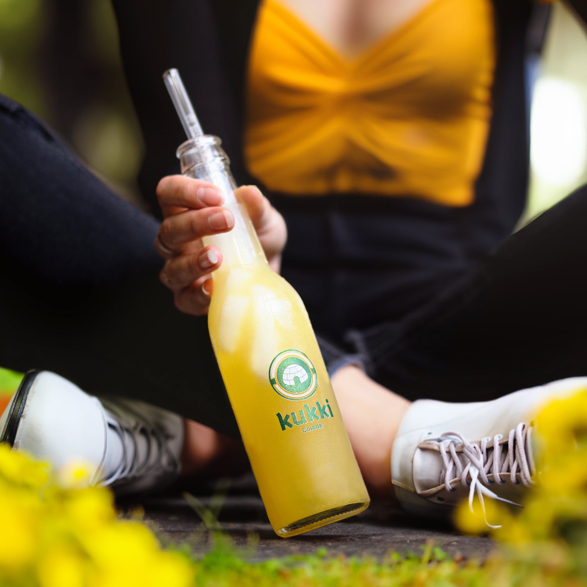 Hand hält gekühlte kukki Cocktail Colada Flasche mit Eiswürfeln und Strohhalm, gelber Deckel, weiße Stiefel im Hintergrund