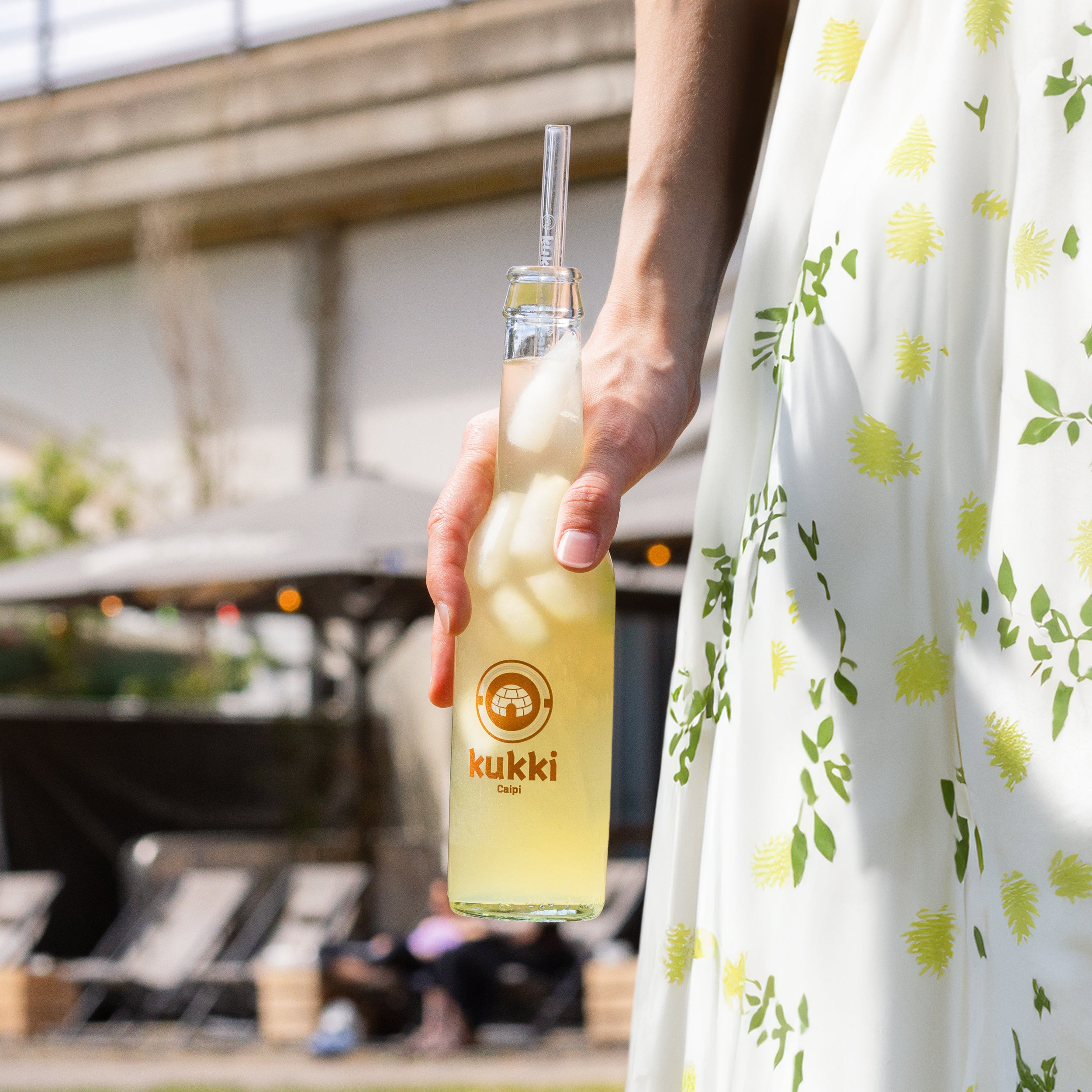 Hand hält eine Glasflasche kukki Cocktail Caipi mit Eis und Strohhalm vor sommerlichem Outdoor-Ambiente.