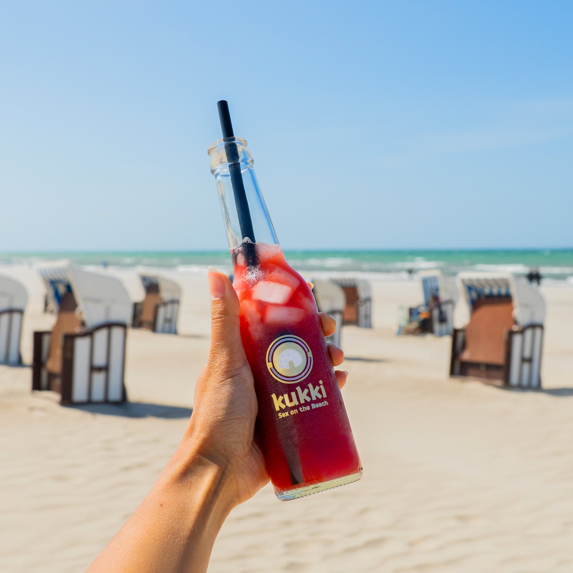 Hand hält Glasflasche mit kukki Cocktail Sex on the Beach und Eiswürfeln vor Strandkulisse mit Stühlen und Meer.