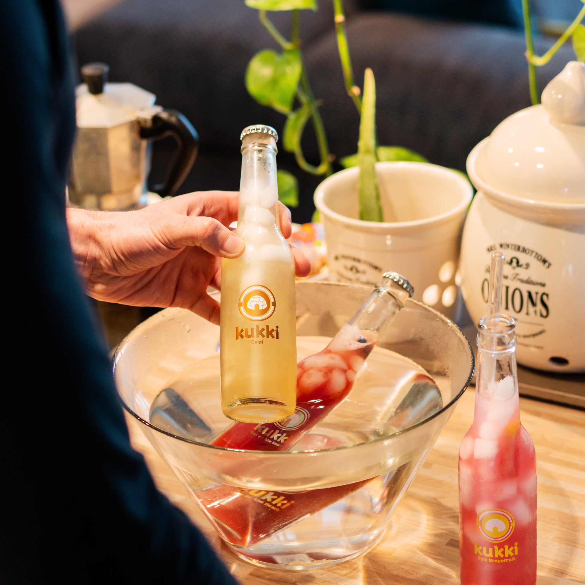 Hand hält kalte Glasflasche kukki Caipi mit gelbem Getränk vor Eis und kukki Pink Grapefruit auf Holztisch.