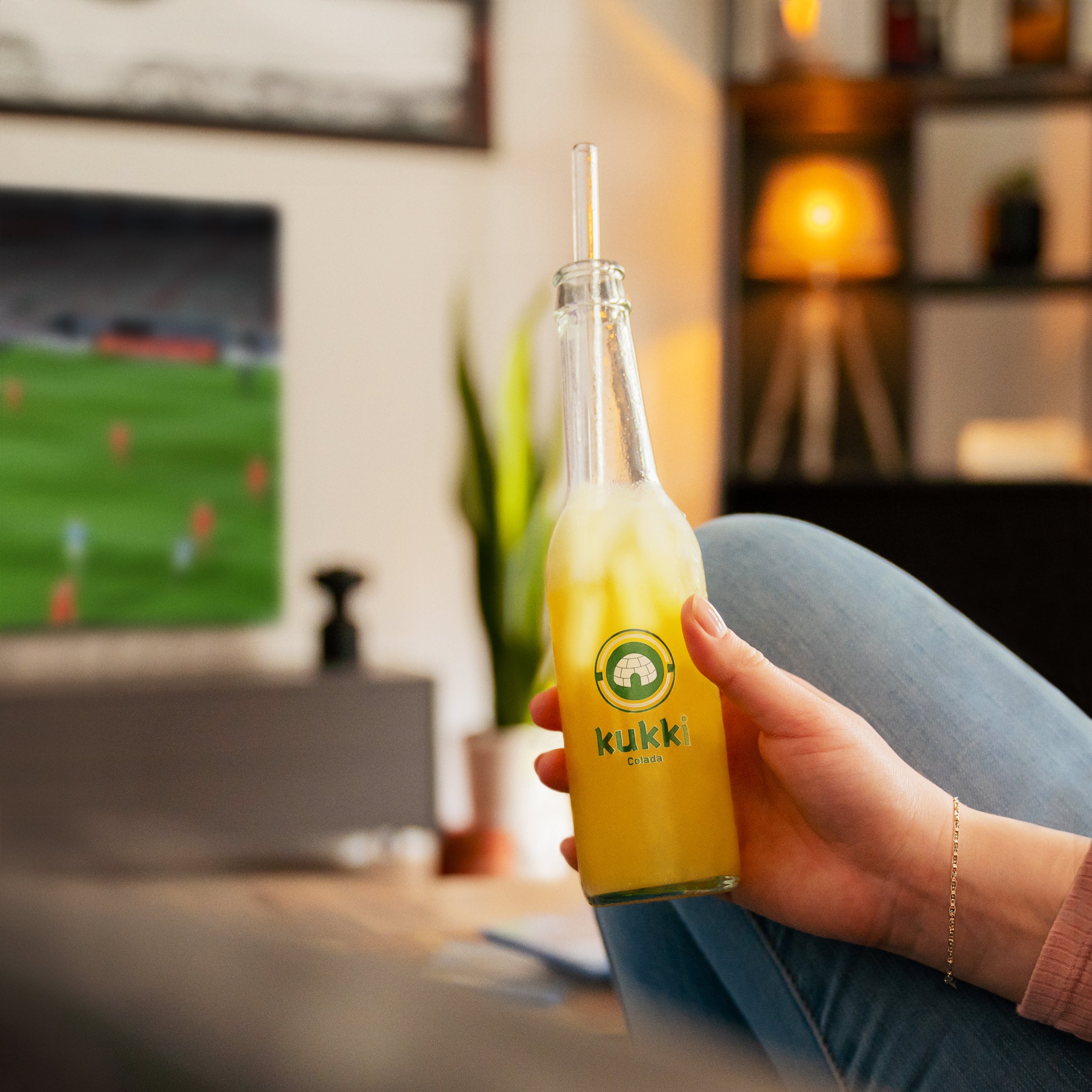 Hand hält kukki Cocktail Colada eisgekühlt im Wohnzimmer mit Fernseher und Pflanze im Hintergrund.