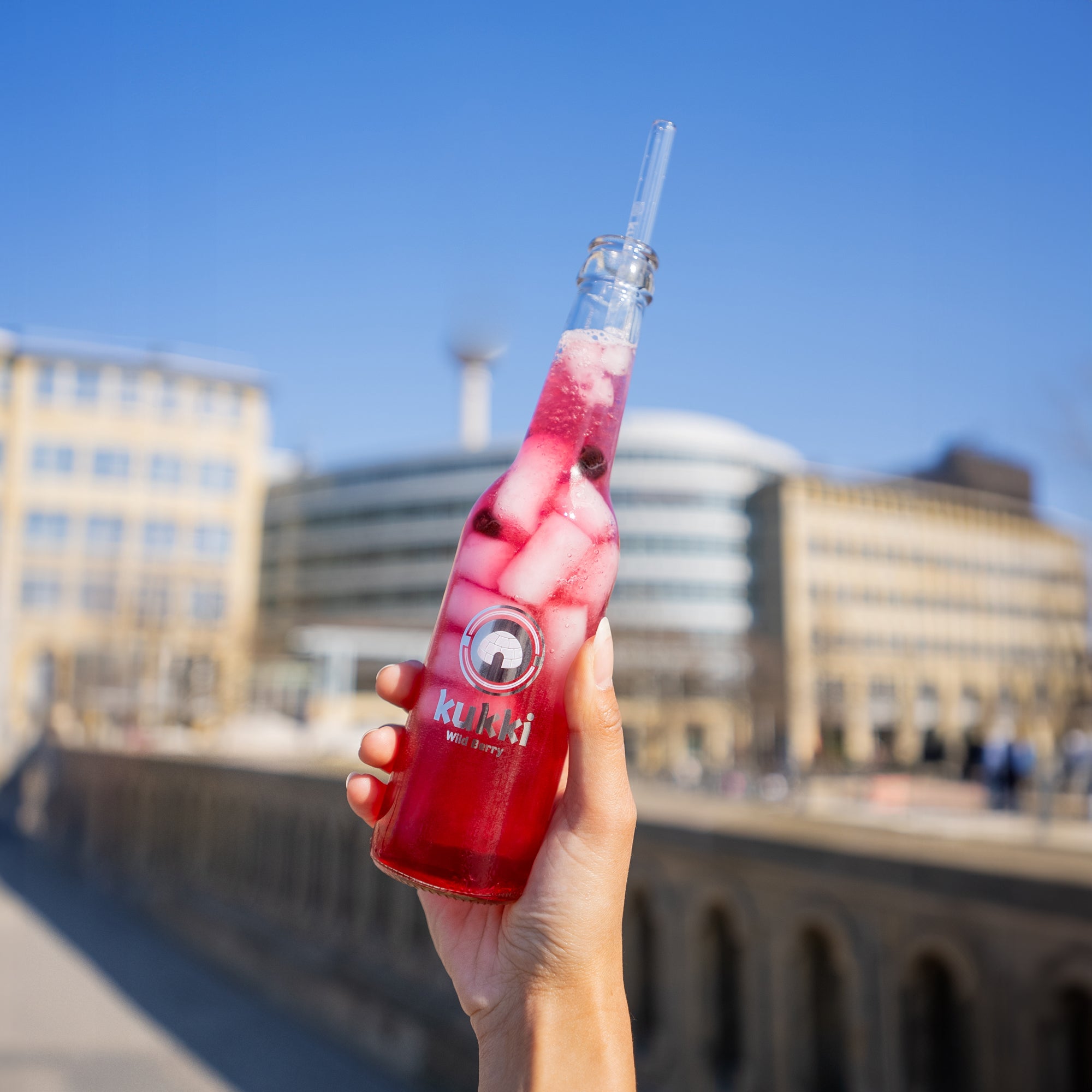 Hand hält eine Glasflasche mit kukki Cocktail Wild Berry, Eiswürfeln und dunklen Beeren vor urbanem Hintergrund.