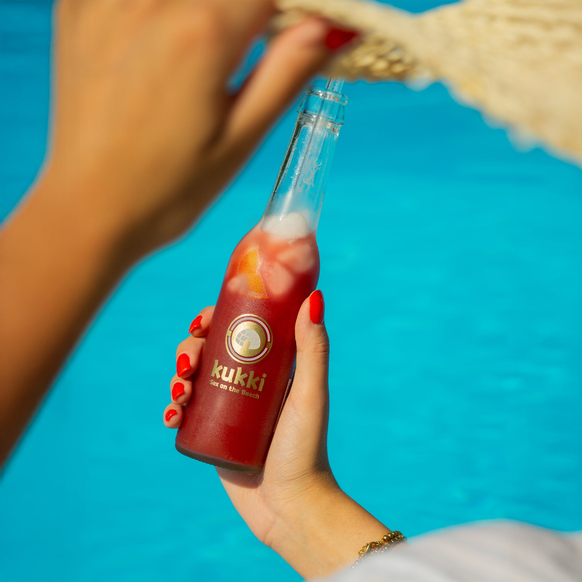 Hand mit rot lackierten Nägeln hält kukki Cocktail Sex on the Beach vor blauem Pool