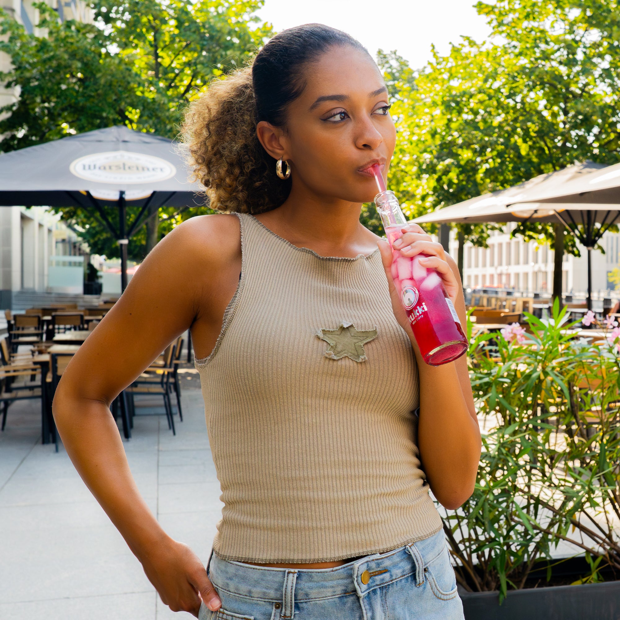 Junge Frau mit lockigen Haaren trinkt pinken eisgekühlten kukki Cocktail aus Glasflasche im sonnigen Café.