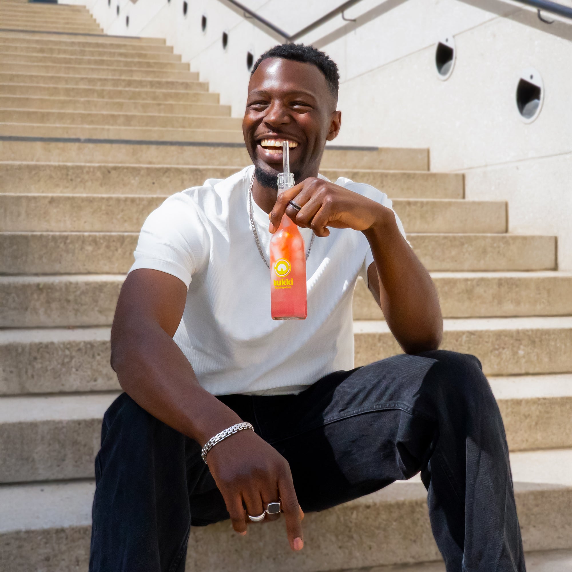 Junger Mann sitzt auf Betonstufen und trinkt kukki Cocktail Pink Grapefruit aus Glasflasche mit Strohhalm