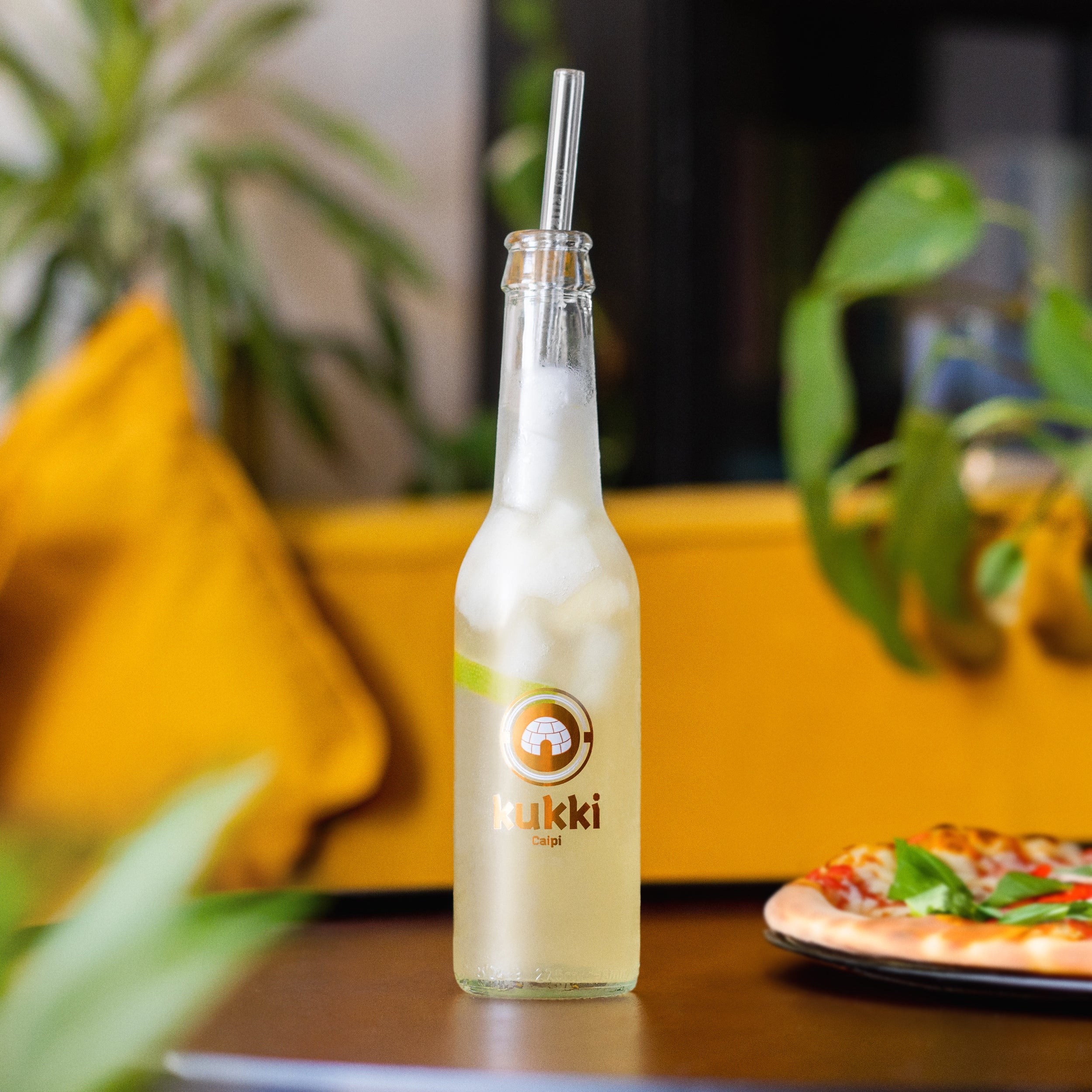 kukki Cocktail Caipi in Glasflasche mit Eis und Metallstrohhalm auf Tisch neben Pizza mit Basilikum
