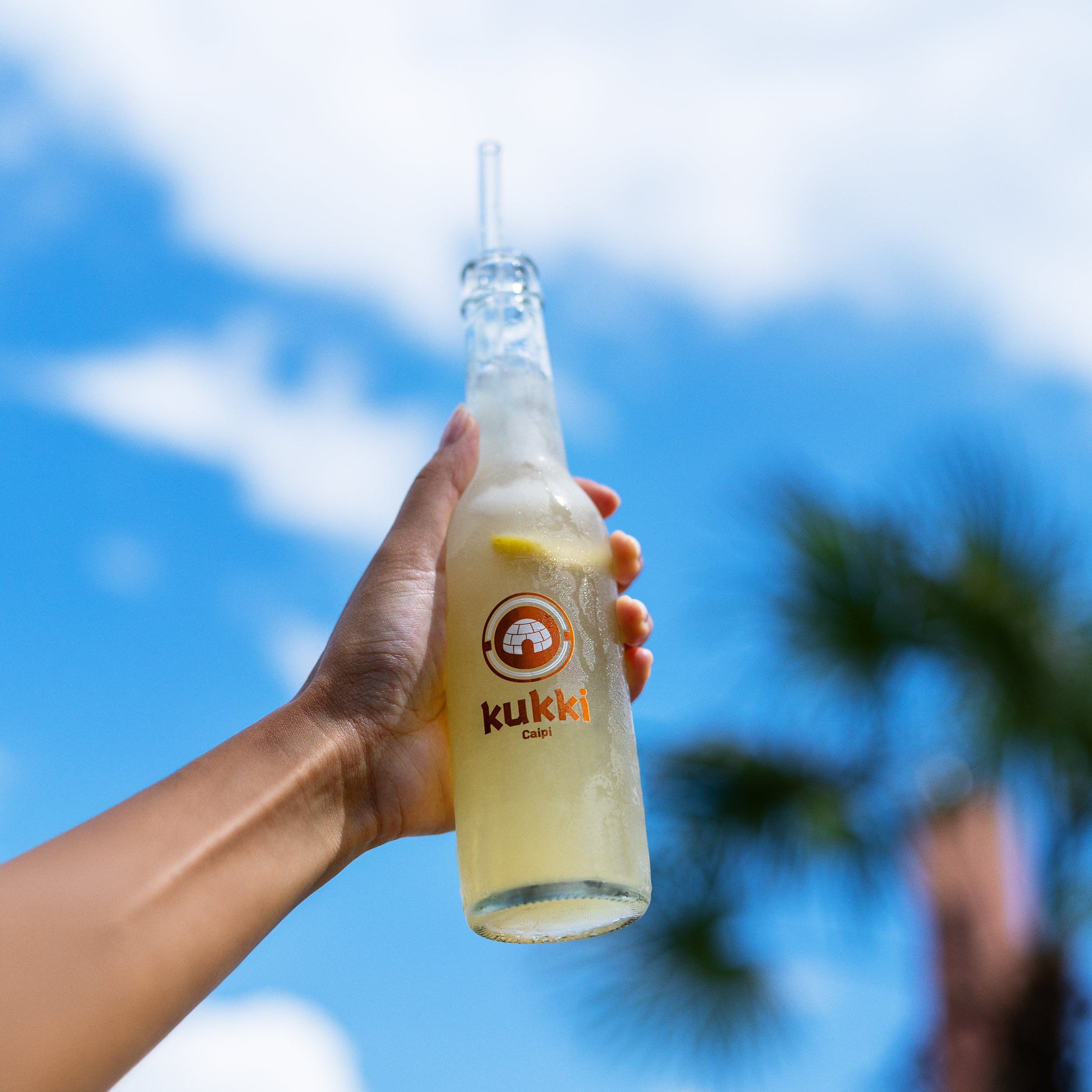 kukki Cocktail Caipi in frostiger Glasflasche mit Zitrusscheibe und Strohhalm vor tropischem Himmel