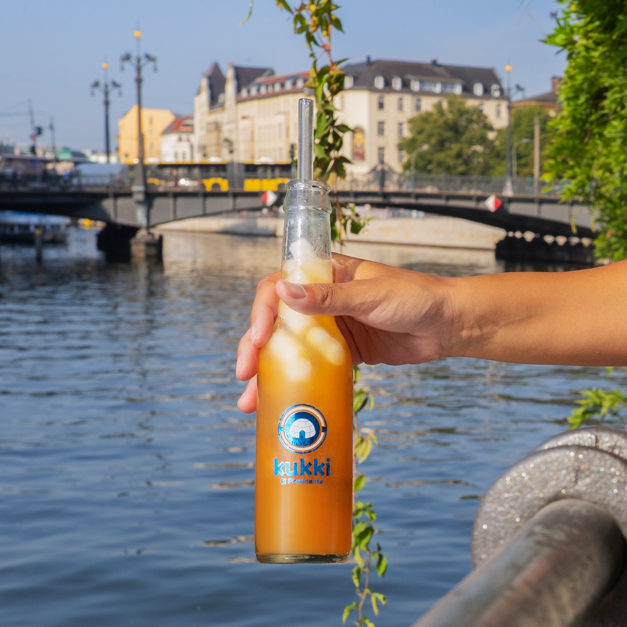 Hand hält eine Glasflasche mit eisgekühltem kukki Cocktail El Presidente vor ruhigem Fluss und historischen Gebäuden.