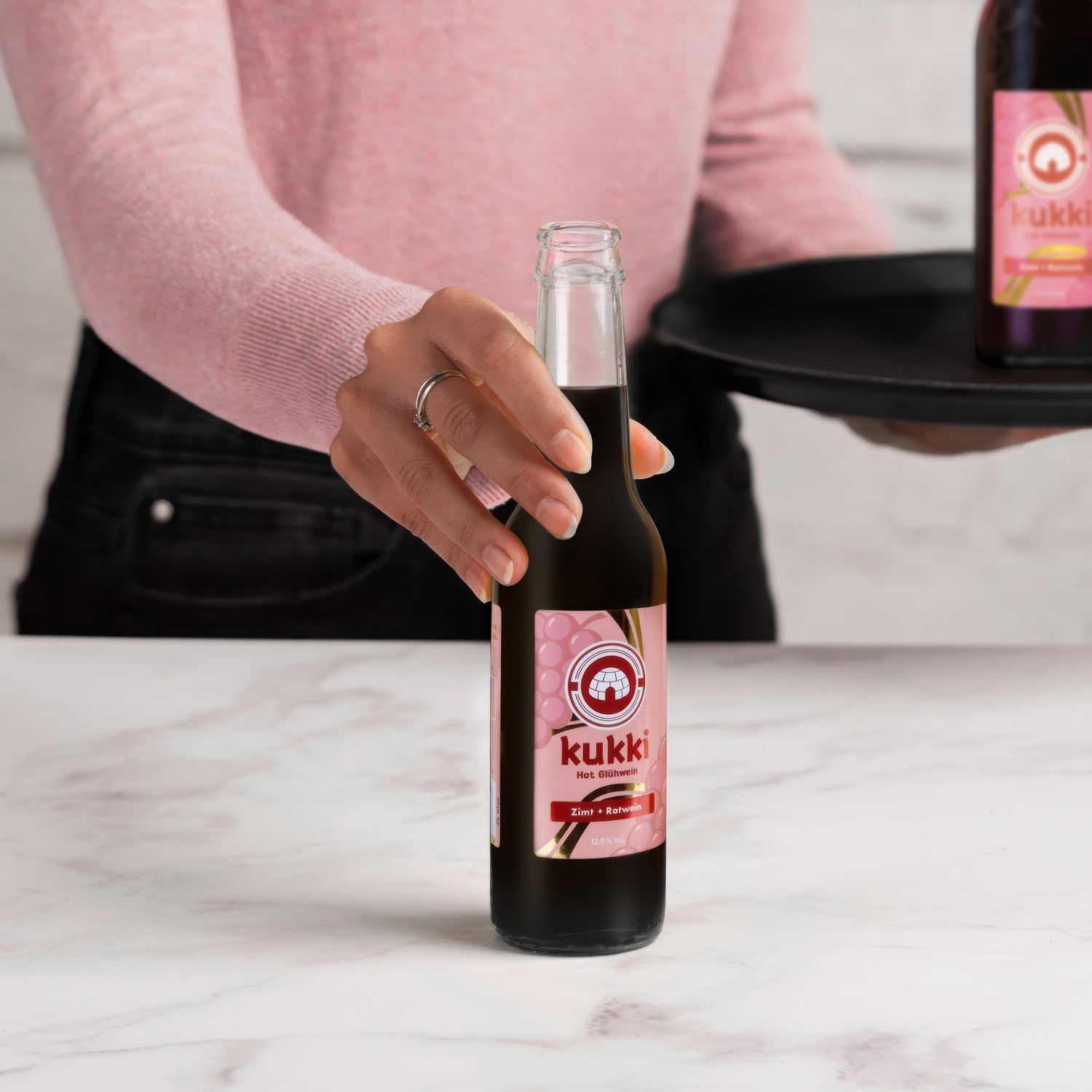 Hand hält eine dunkle Glasflasche kukki Cocktail Glühwein mit Zimt und Rotwein, 12% Vol, auf weißem Marmor.