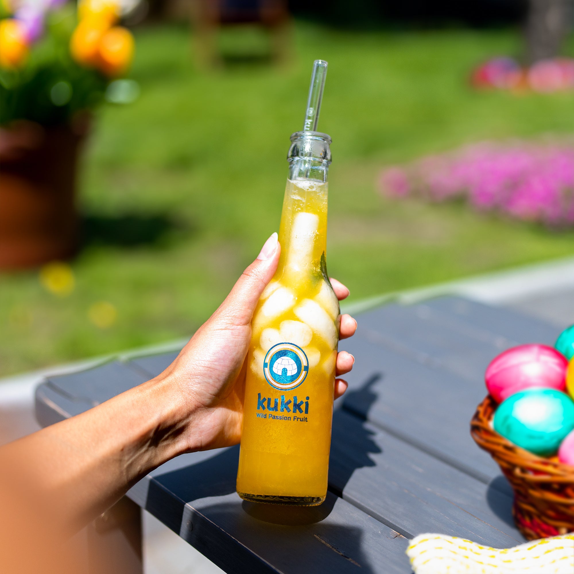 kukki Cocktail Glasflasche Wild Passion Fruit mit Eis und Trinkhalm, Ostereier im Korb auf Tisch im Garten