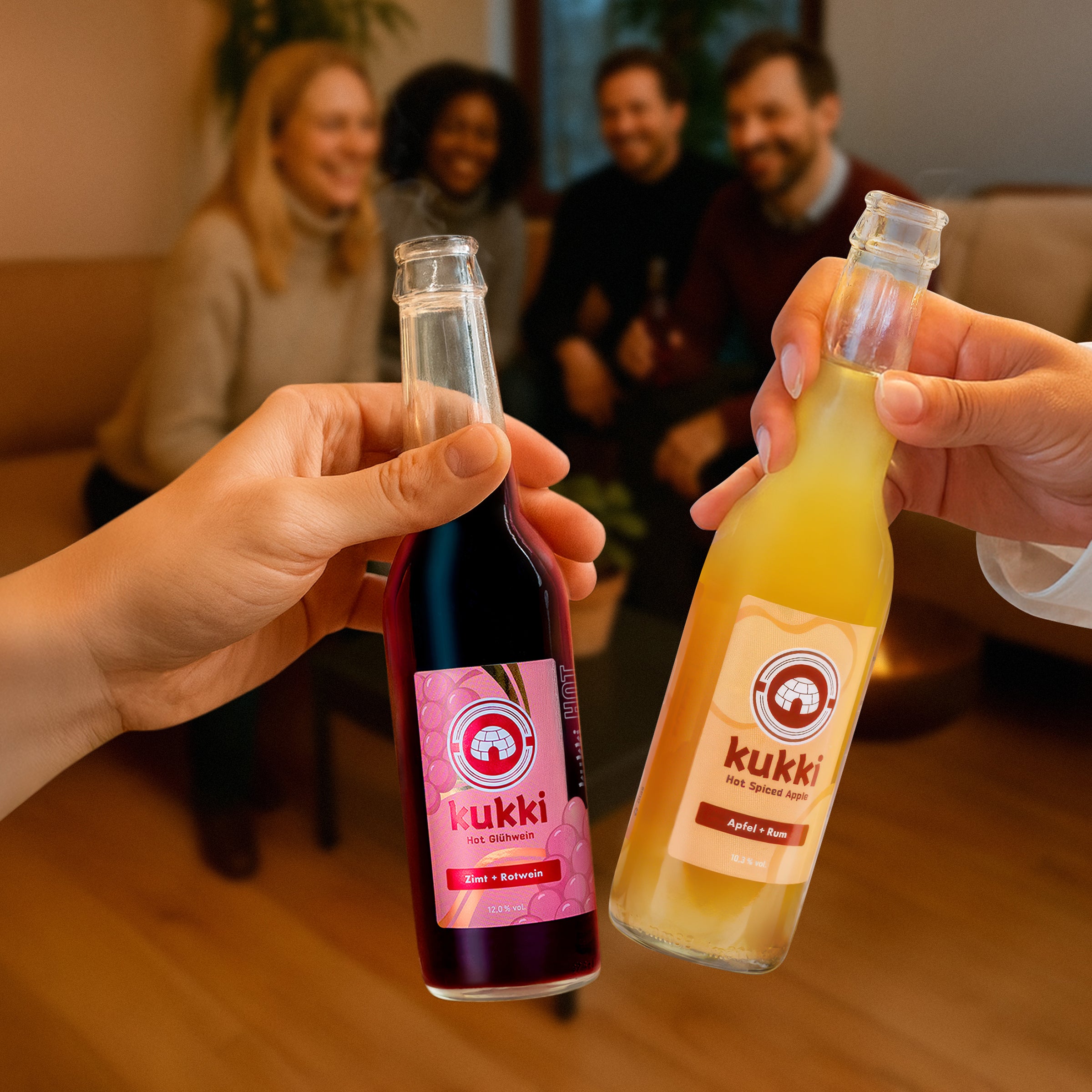 Zwei Hände halten Glasflaschen mit kukki Hot Glühwein Zimt Rotwein und Hot Spiced Apple vor vier lächelnden Personen auf einer Couch in gemütlicher Umgebung.