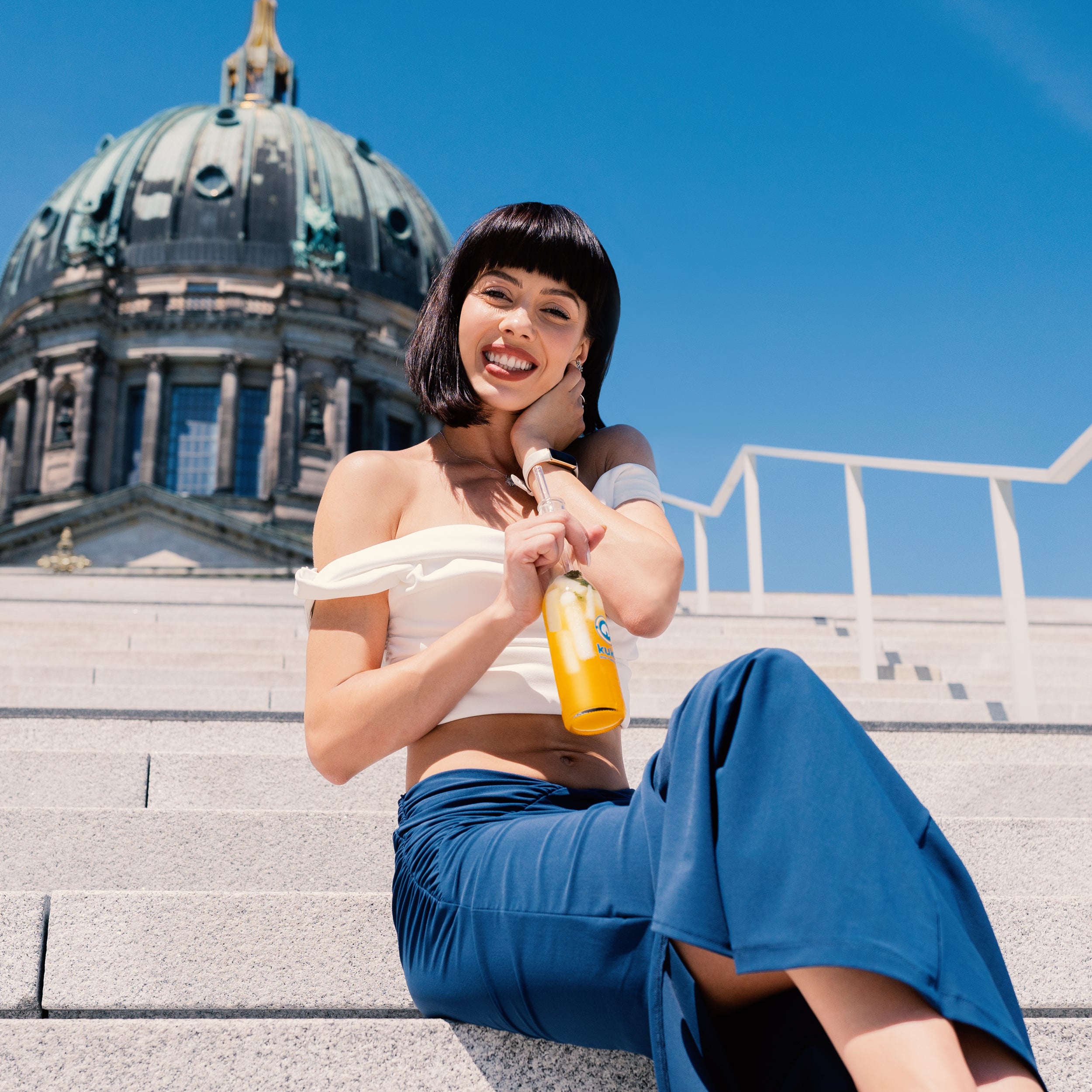Lächelnde Frau in weißem schulterfreiem Oberteil mit kukki Cocktail Caipi auf Treppe.
