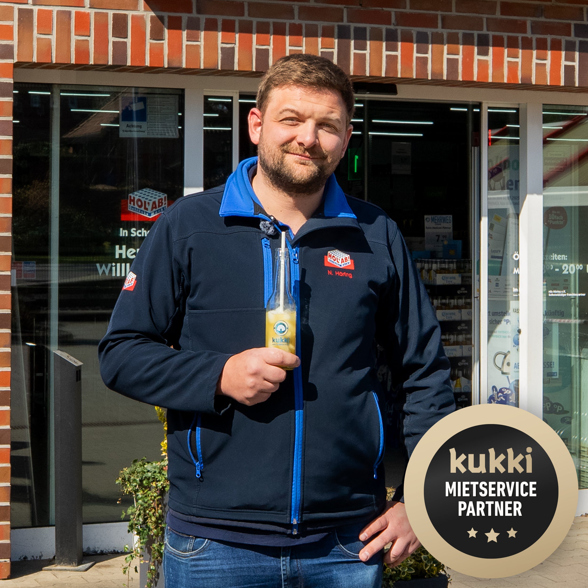 Mann in navyblauer holab-Jacke mit Stickerei hält kukki Cocktail vor Backsteinladen mit Mehrweg-Schaufenster.