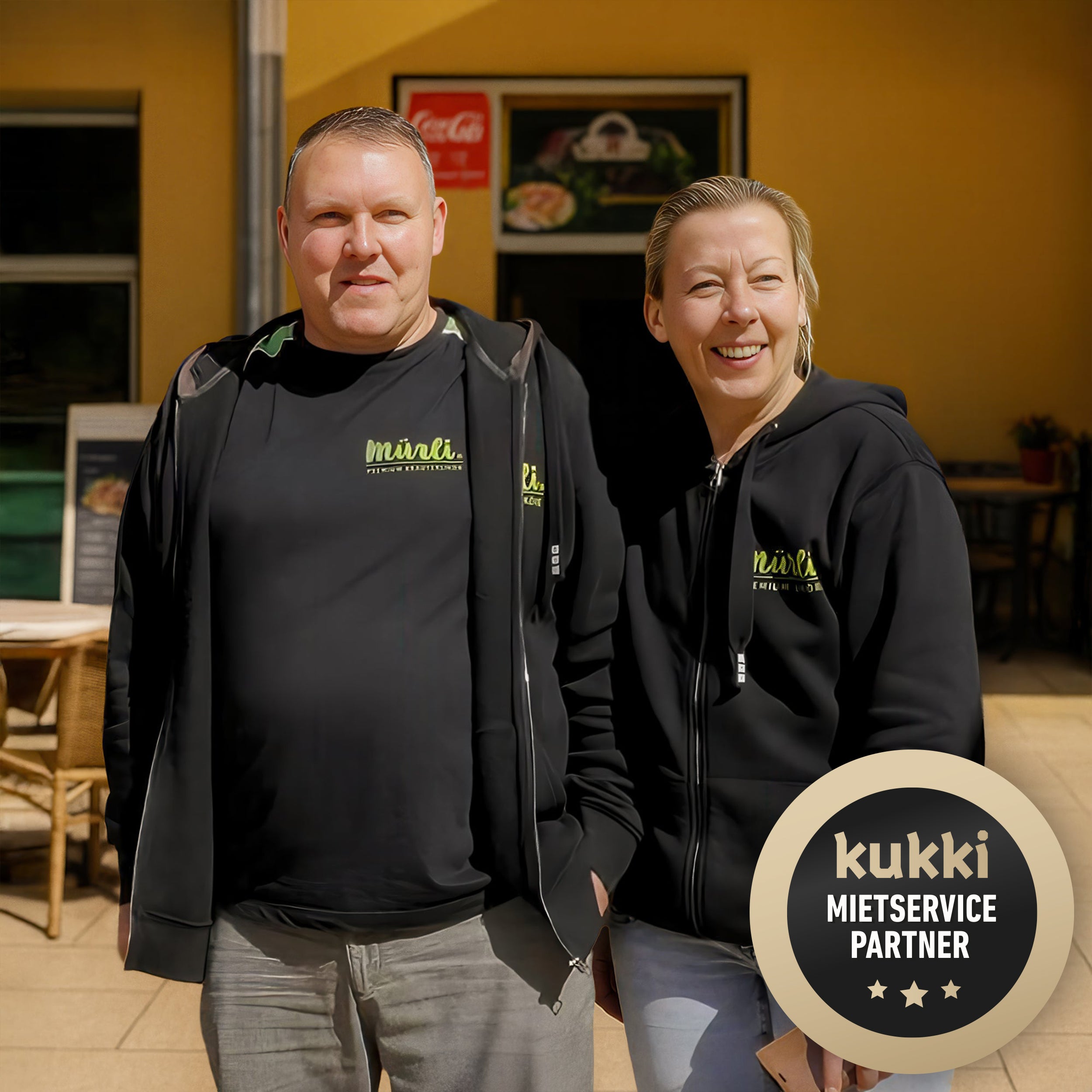 Zwei lächelnde Personen in schwarzer Mürli-Kleidung vor gelber Wand mit kukki Mietservice Abzeichen.